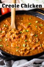 A large black cast-iron skillet filled with a chicken creole sauce with a wooden spoon inserted into the middle.