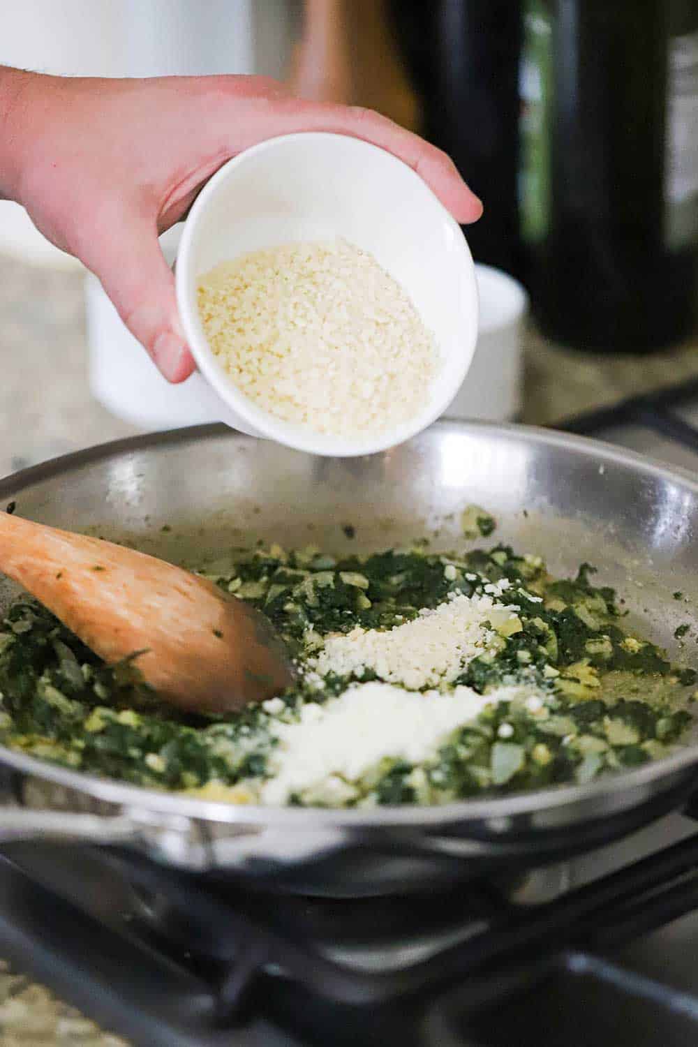 A hand sprinkling Panko bread crumbs from a small white bowl into a skillet filled sautéd spinach and onions. 