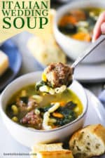 A hand holding a soup spoon with a meatball, some pasta, and spinach on it and over a bowl of the soup.