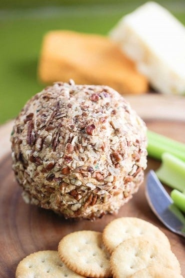 A close-up view of a cheese ball that is encased in chopped pecans sitting on a board next to crakers and celery sticks. 