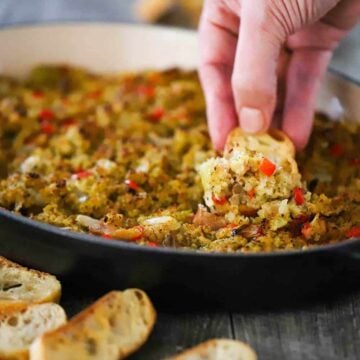 A hand using a slice of toasted baguette to scoop up a helping of baked clam dip.