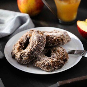 Three apple cider doughnuts on a white plate with an apple and apple cider next to it.
