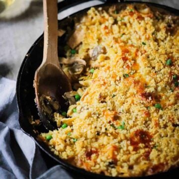 A large black cast-iron skillet filled with tuna casserole with a serving missing from the pan and a wooden spoon inserted in the side of the pan.