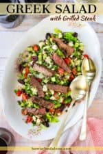 A large oval platter filled with a Greek salad and grilled steak with two large gold spoons on the side.
