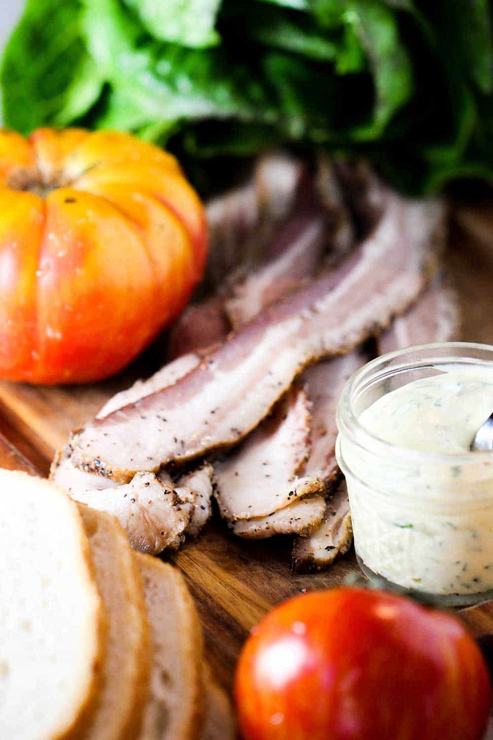 An heirloom tomato, basil, bread slices, bacon and aioli on a wooden cutting board. 