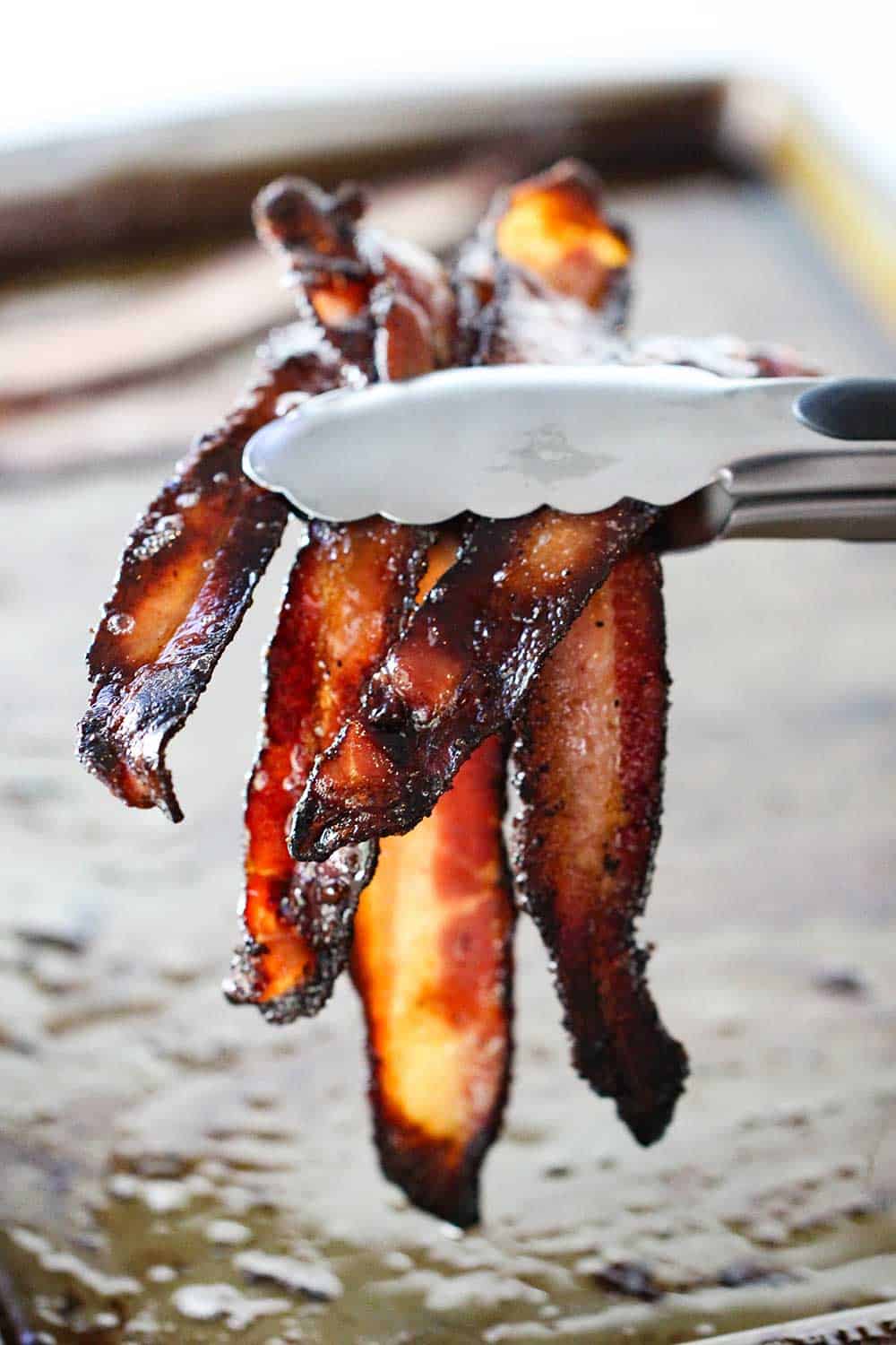 A pair of tongs holding up 5 or 6 slices of cooked black pepper bacon over a baking sheet. 