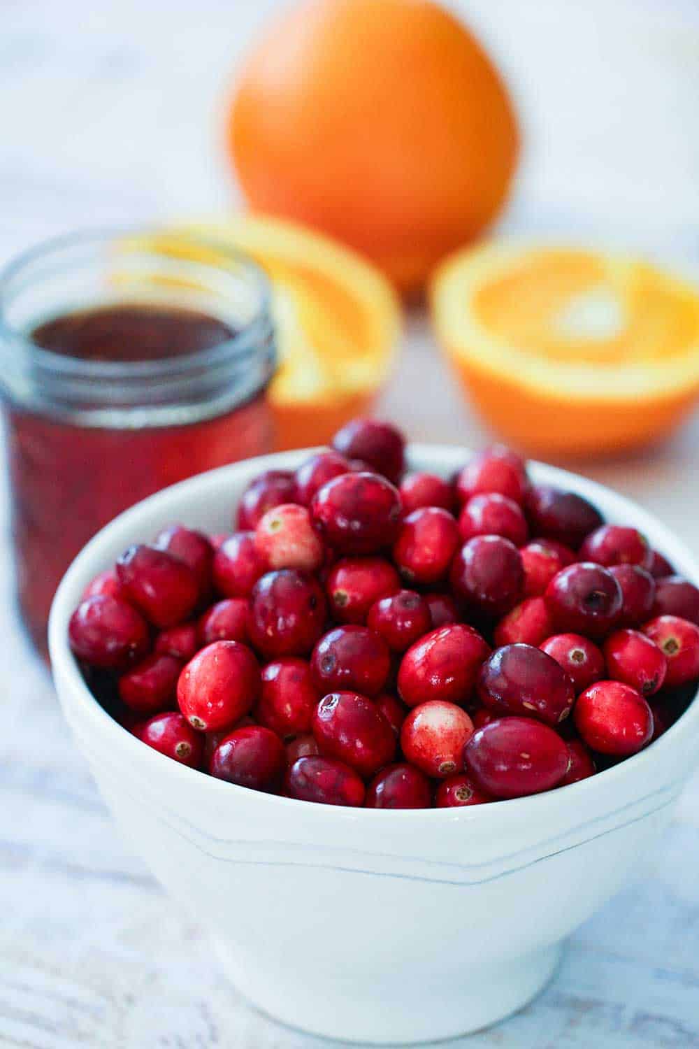 A white bowl holding fresh cranberries next to maple syrup and an orange. 