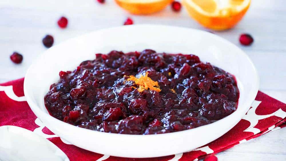A white bowl holding a serving of Maple Orange Cranberry Sauce with a sliced orange next to it. 