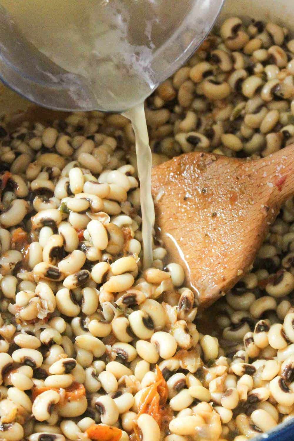 Chicken stock pouring into a pot of cooking black-eyed peas with tomatoes