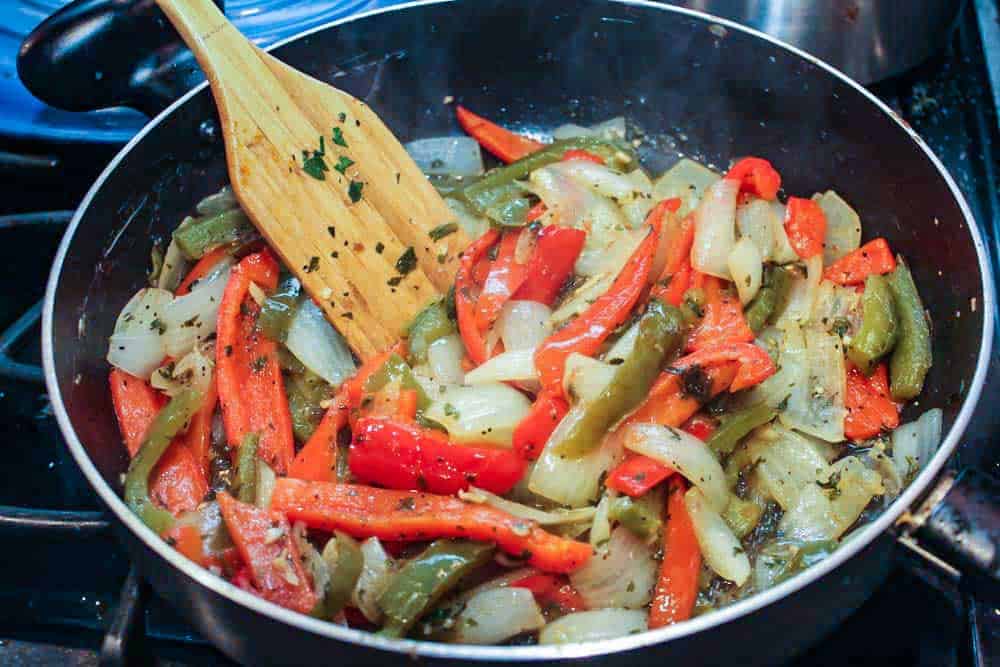 Red and green bells peppers and sliced onion and herbs cooking in a large skillet with a wooden spoon. 