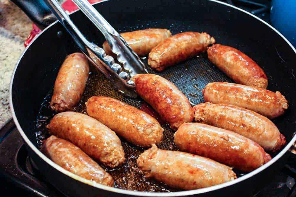 Hot Italian sausages cooking a in large skillet with a pair of tongs moving them around. 