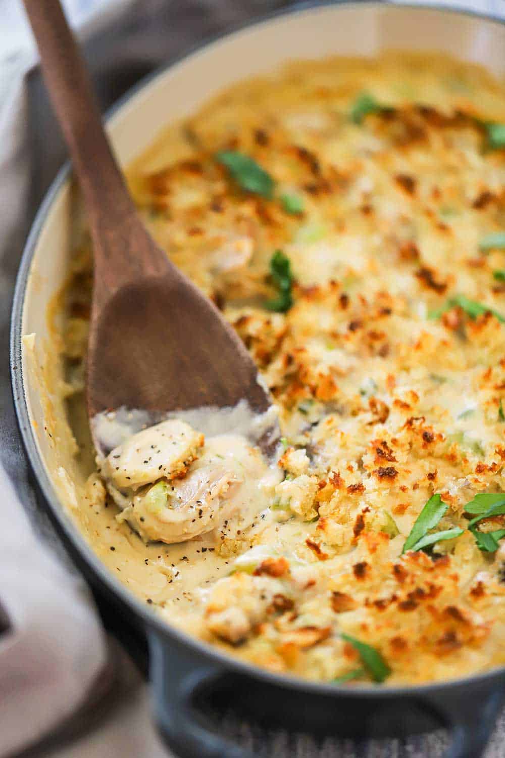 A wooden spoon sitting in casserole dish filled with cooked oyster casserole, with one serving missing from the dish. 