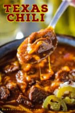 A spoon lifting a helping of Texas-style chili from a bowl of the chili with cheese dripping off the sides.