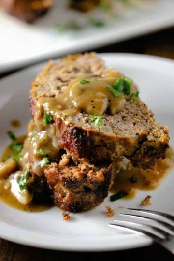 Two slices of best-ever meatloaf sitting on top of each other on a white plate with mushroom gravy on top.