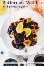A stack of buttermilk waffles topped with a blueberry sauce and a slice or orange all on a white plate with a fork next to it.