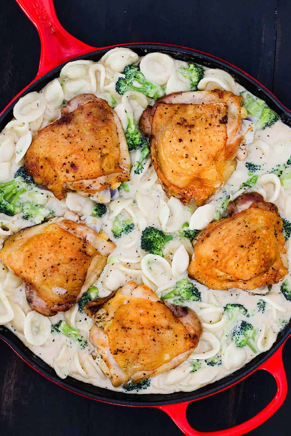 An overhead view of a large red skillet holding seared chicken thighs in a broccoli and pasta alfredo sauce. 