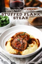 A straight-on view of a white dinner bowl filled with a serving of creamy polenta topped with two slices of stuffed flank steak and topped with a wine reduction sauce.