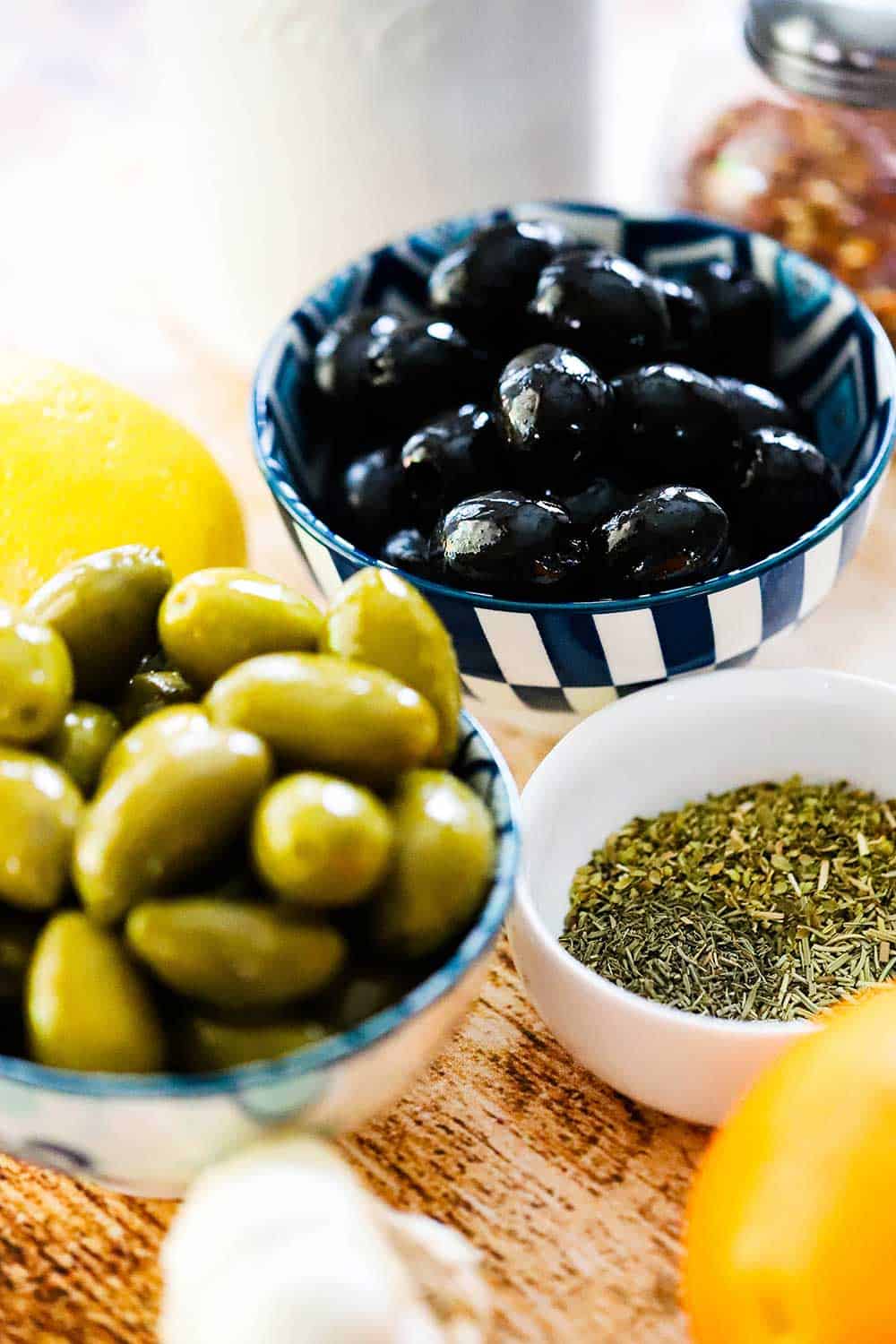 One small bowl holding green olives and another bowl holding black olives surround by dried herbs, an orange, and an olive oil dispenser. 