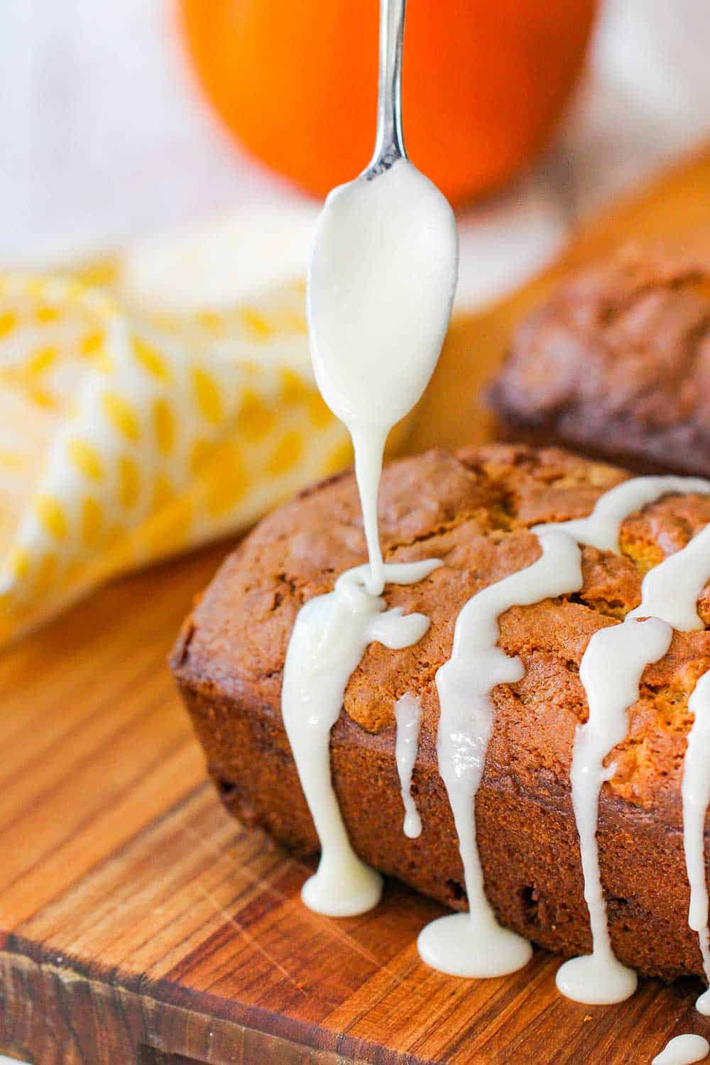 A spoon drizzling a sugar glazed over freshly baked pumpkin bread on a cutting board. 