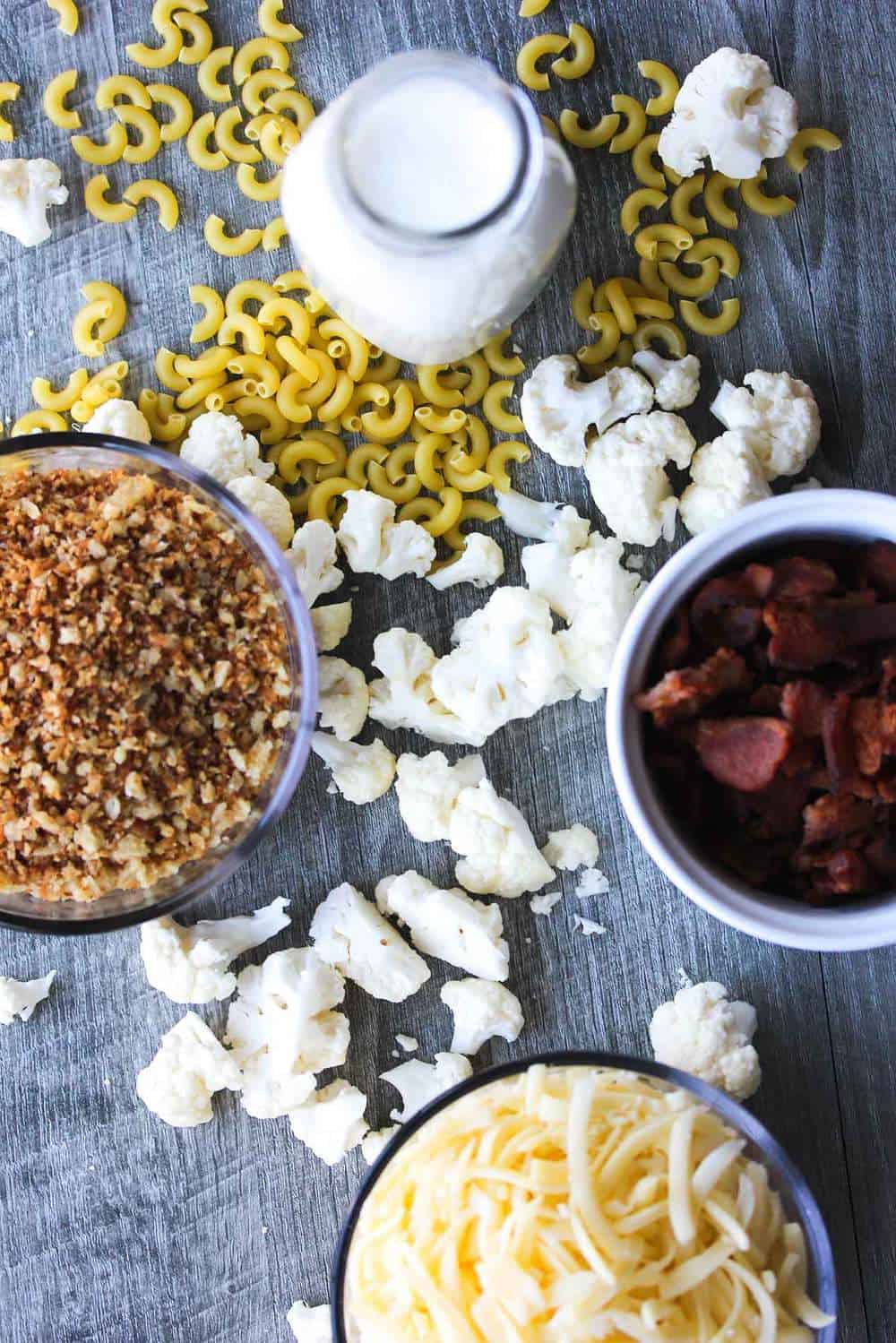 A spread of ingredients including uncooked elbow macaroni, a jug of milk, a bowl of shredded cheese, a bowl of crumbled cooked bacon, and a bowl of toasted breadcrumbs. 