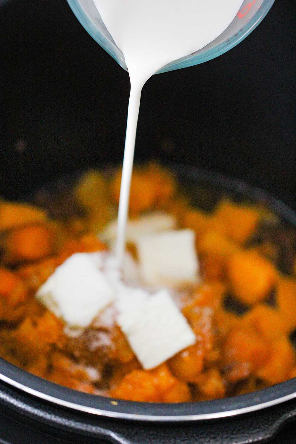 A hand pouring heavy cream into a pot of steamed butternut squash and butter. 