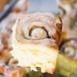 A hand holding a spatula with a homemade cinnamon roll on it over a pan of the rolls.