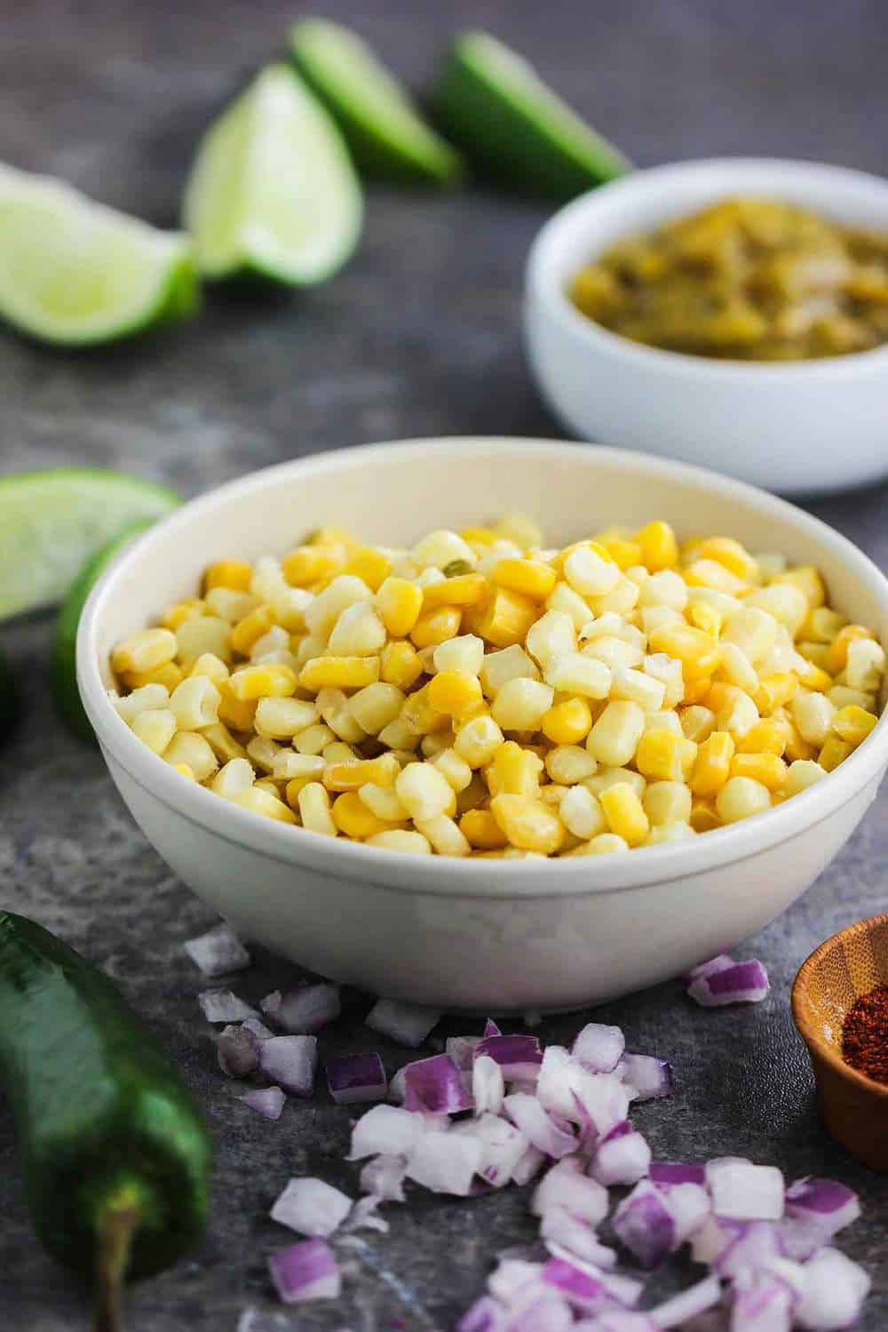 A medium-sized white bowl filled with fresh corn kernels with scattered chopped red onion around the base of the bowl
