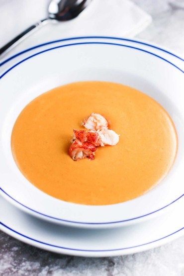 Lobster bisque in a white bowl with blue rim next to a spoon and white napkin