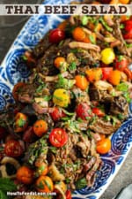 A colorful salad consisting of grilled strips of skirt steak, cherry tomatoes, shallots, and a Thai herb dressing all on an oval blue platter.