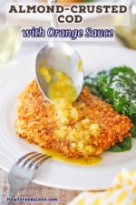 A dinner spoon being used to drizzle an orange shallot sauce over an almond crusted fillet of cod next to a spinach salad.