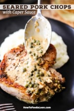 A person using a silver spoon to drizzle on a creamy caper sauce over a seared pork chop on a black plate with mashed potatoes.