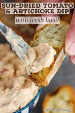 A person using a small butter knife to spread sun-dried tomato and artichoke dip onto a toasted slice of a baguette.