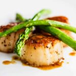 A white dinner plate filled with seared scallops topped with sautéed asparagus topped with a brown sauce.