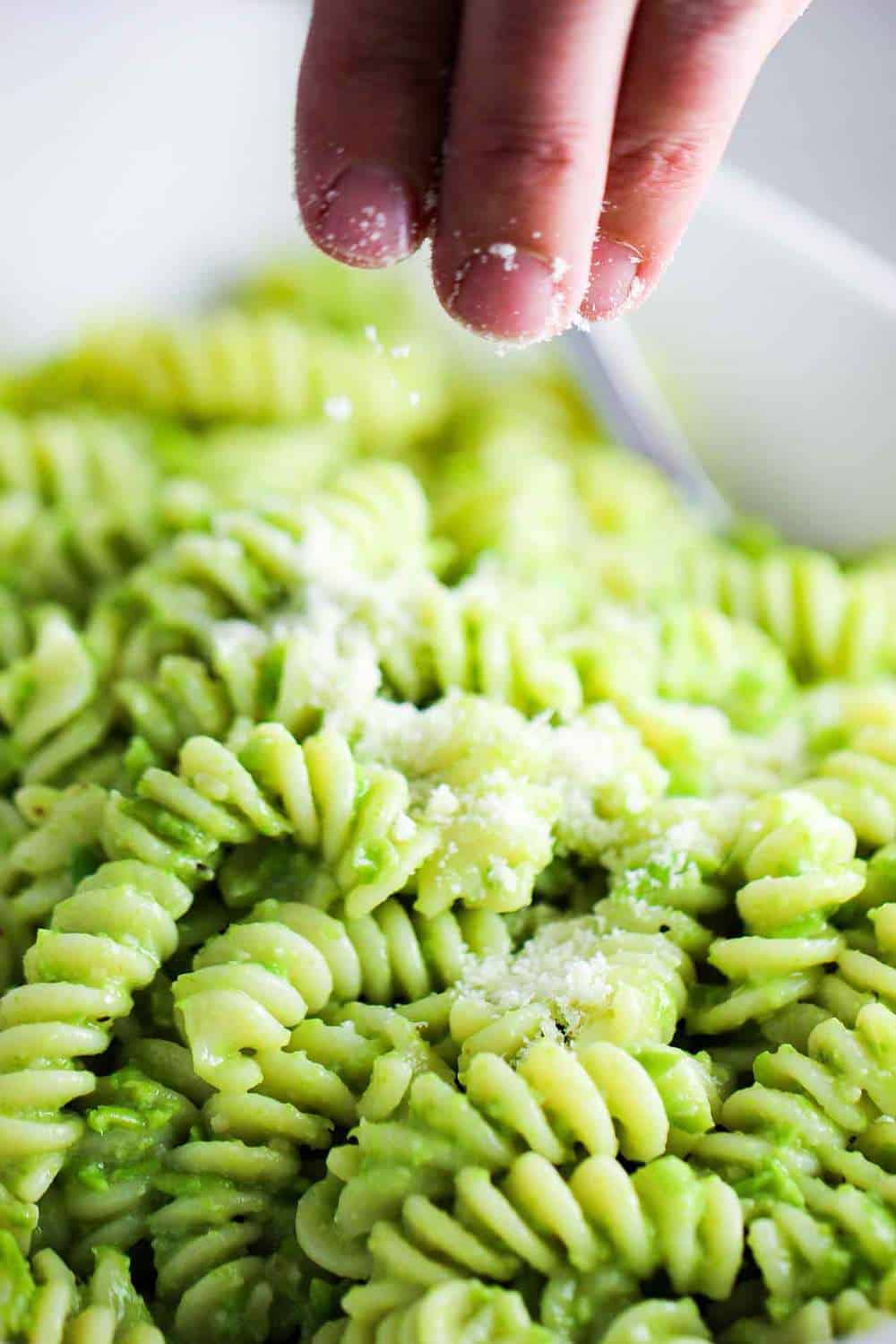 Fingers sprinkling grated Parmesan cheese over a bowl of pea pesto pasta
