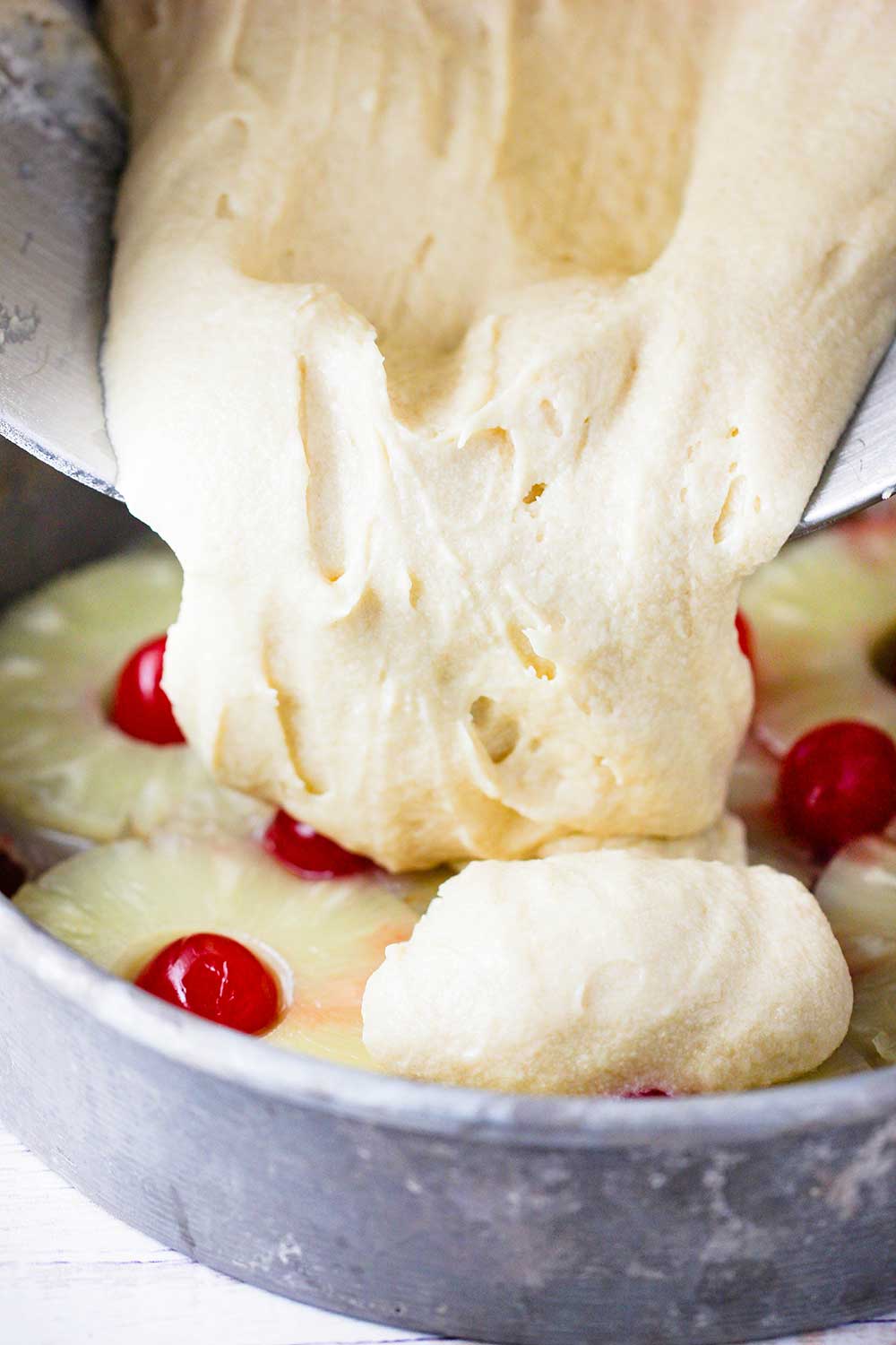 Yellow butter cake batter being poured into a cake pan with pineapples and cherries.
