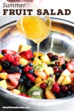 A small glass pitcher of being used to pour an orange honey dressing over cut fruit in a metal bowl.