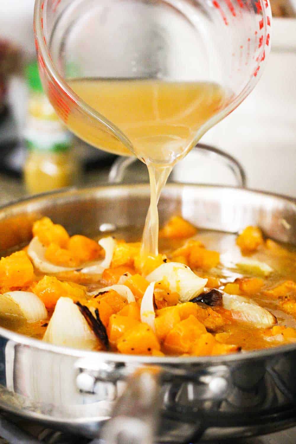 A large measuring cup pouring broth into a skillet of vegetables for butternut squash soup. 
