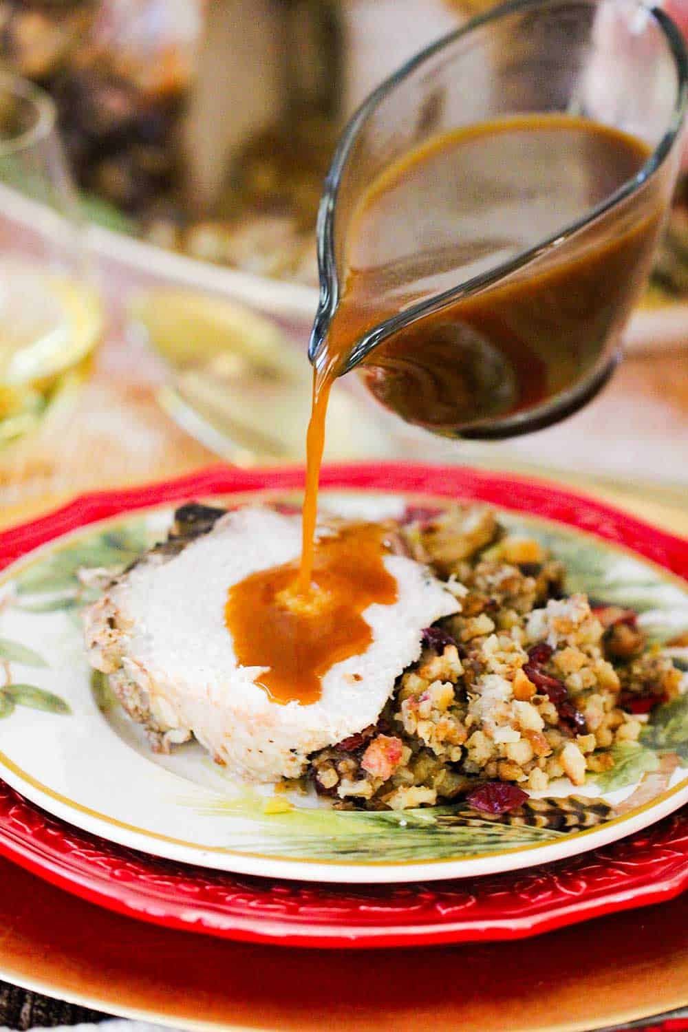A hand pouring sauce from a gravy boat onto a pork chop next to stuffing on a red plate. 