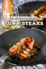 A straight-on view of two black dinner plates side-by-side both filled with Mediterranean tuna steaks with a glass of white wine nearby.