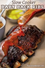 A person pouring homemade barbecue sauce from a clear glass bottle onto a rack of slow cooker baby ribs that have been fully cooked.