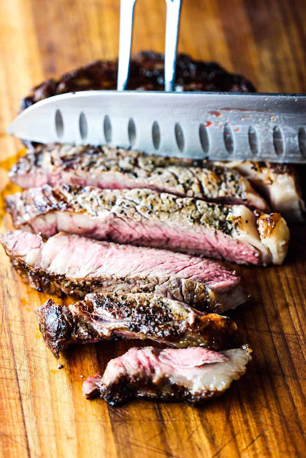 Cooked ribeye steak being sliced by a chef's knife on a wooden cutting board