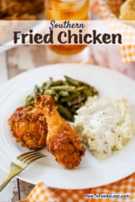 A white dinner plate filled with pieces of Southern fried chicken, a pile of mashed potatoes with cream gravy, and a helping of green beans.
