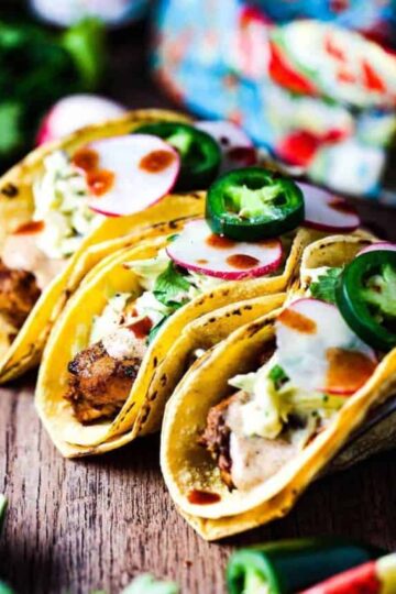 Three mahi mahi tacos sitting on a taco holder topped with sliced radishes and fresh jalapenos.