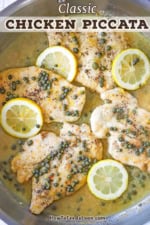 An overhead view of a large stainless steel skillet filled with five seared chicken cutlets that are resting in a lemon, butter, and capers sauce with lemon slices scattered around.