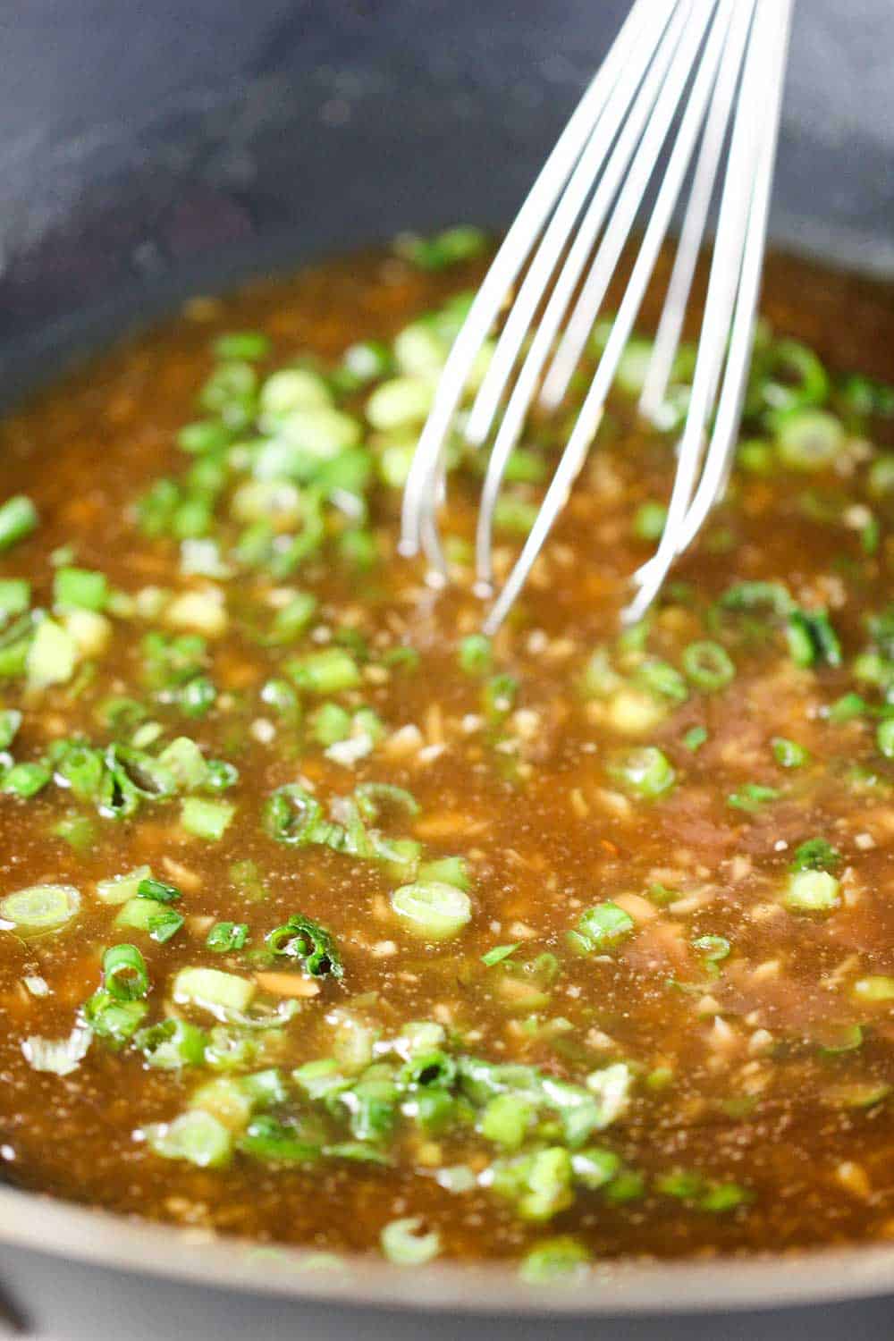 A wok filled with sweet and sour sauce with scallions and a whisk.