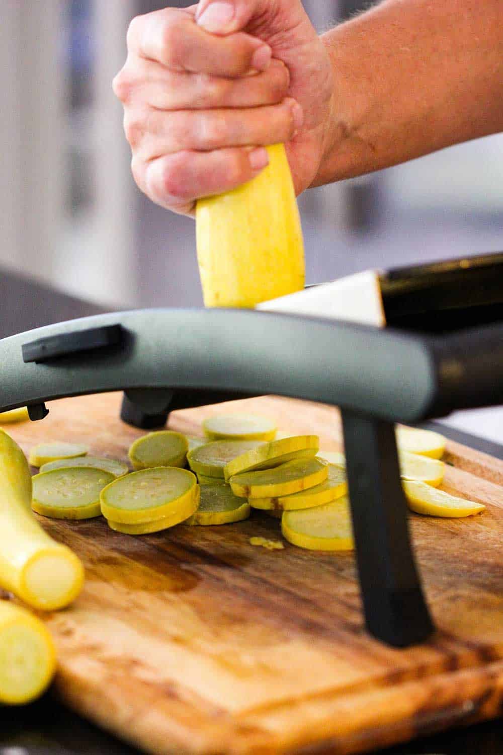 A hand slicing a yellow squash on a mandolin over a cutting board.