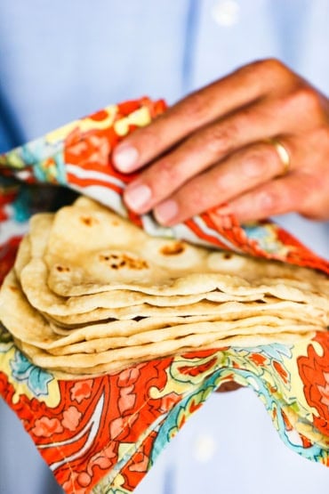 A person pulling back a festive-color napkin to reveal a stack of homemade flour tortilla