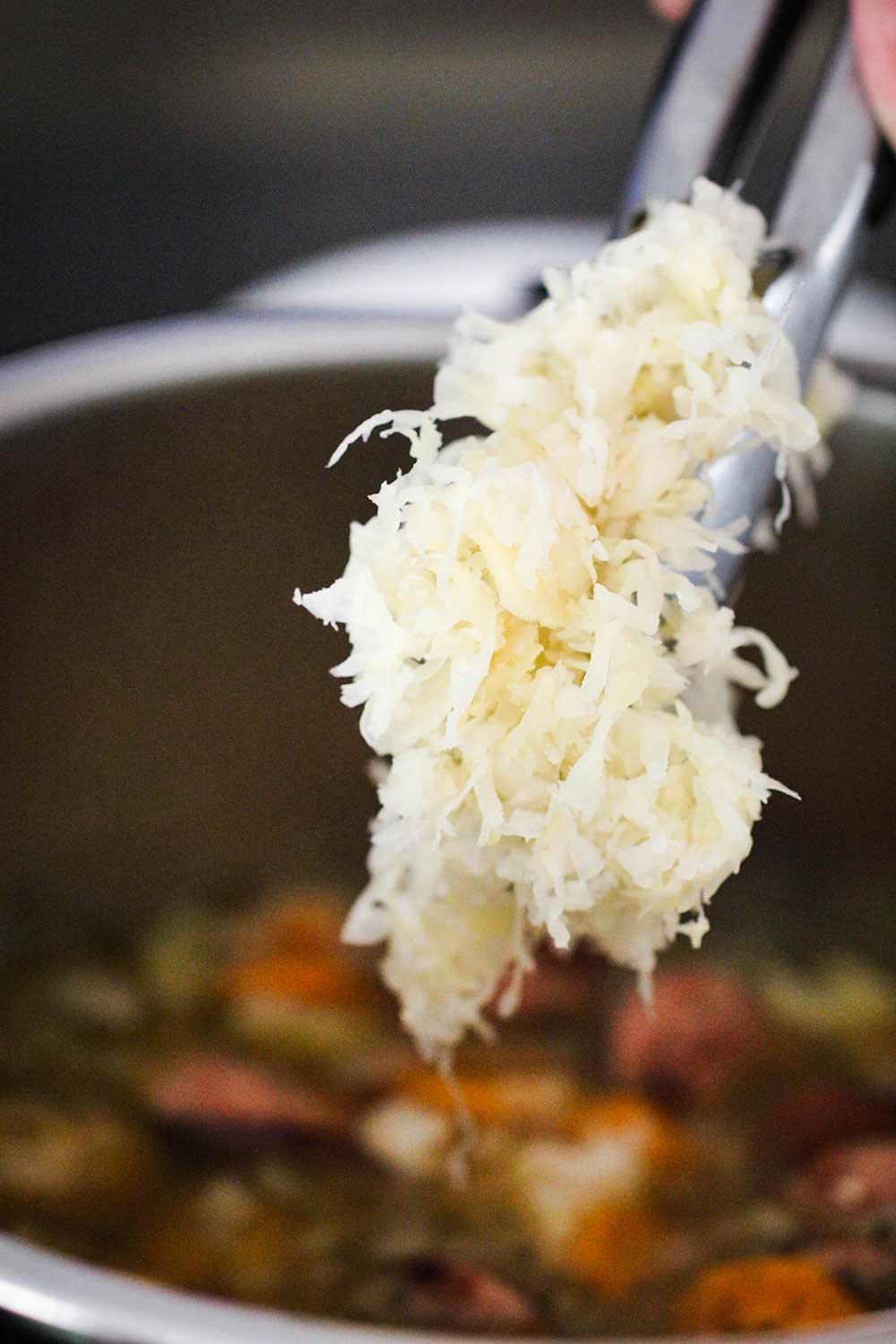 A pair of tongs getting ready to lower sauerkraut into a pot of smoked sausage stew. 