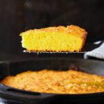 A slice of homemade cornbread being raised out of a skillet with a spatula.