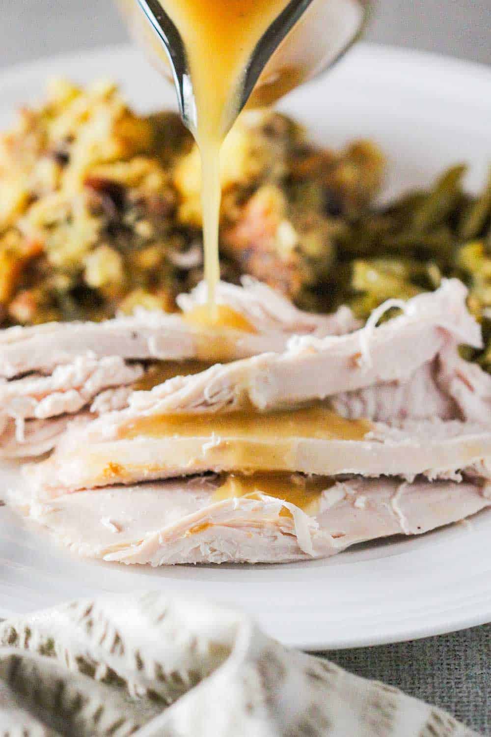 Gravy being poured out of a gravy boat onto sliced turkey on a plate with dressing and beans. 